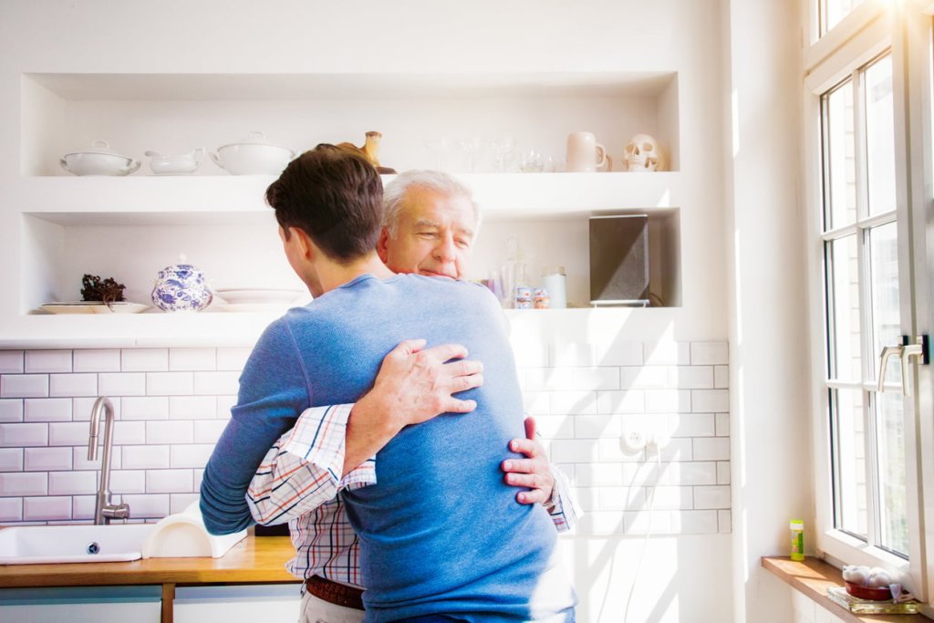 Senior man embraces mature son in sunny kitchen